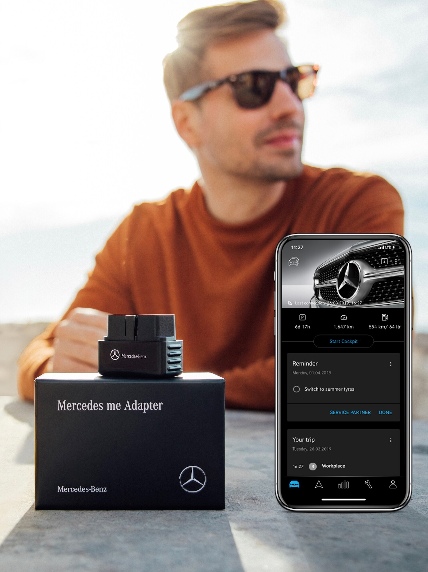 Un hombre con gafas de sol se encuentra sentado frente a una mesa en la que se ve en un primer plano el Mercedes me Adapter y un smartphone con la Mercedes me Adapter App abierta.
