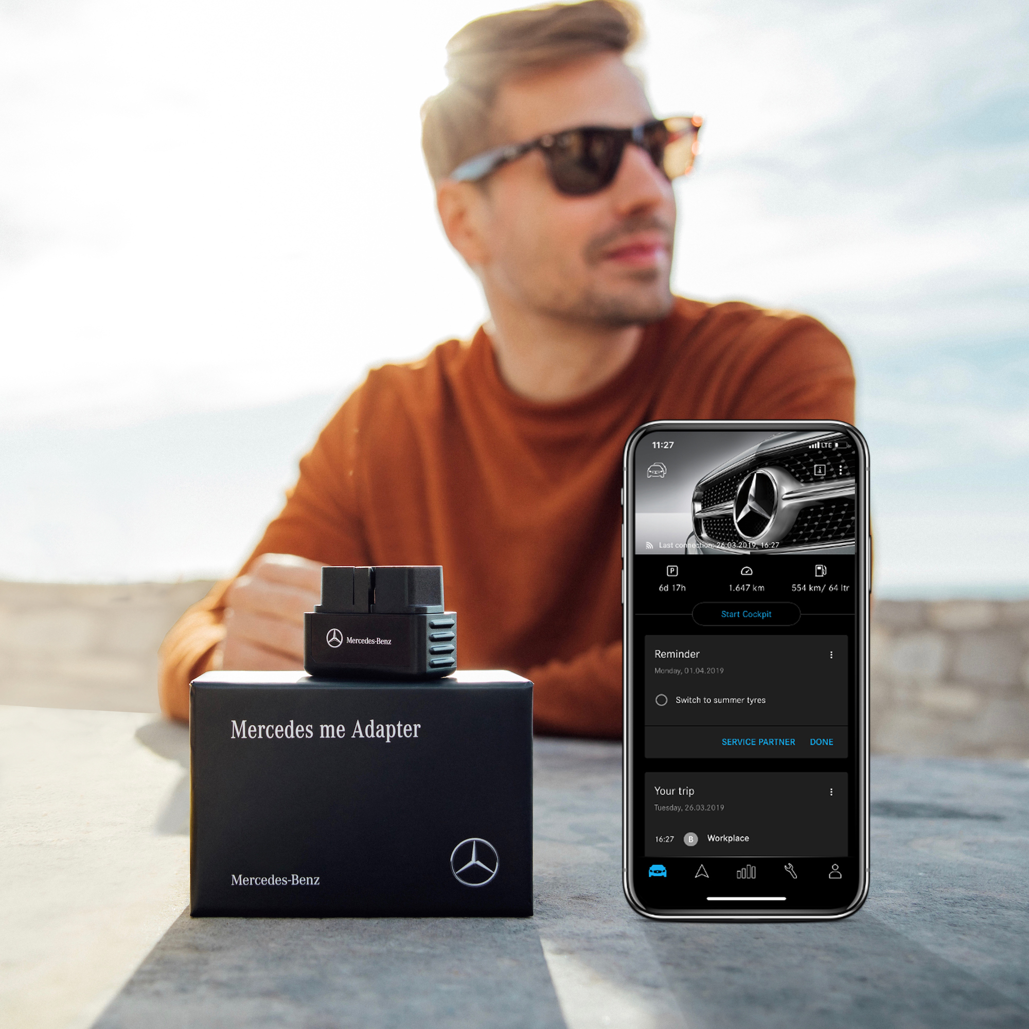 Detección de accidentes con el Mercedes me Adapter. | Mercedes-Benz Un hombre con gafas de sol se encuentra sentado frente a una mesa en la que se ve en un primer plano el Mercedes me Adapter y un smartphone con la Mercedes me Adapter App abierta.