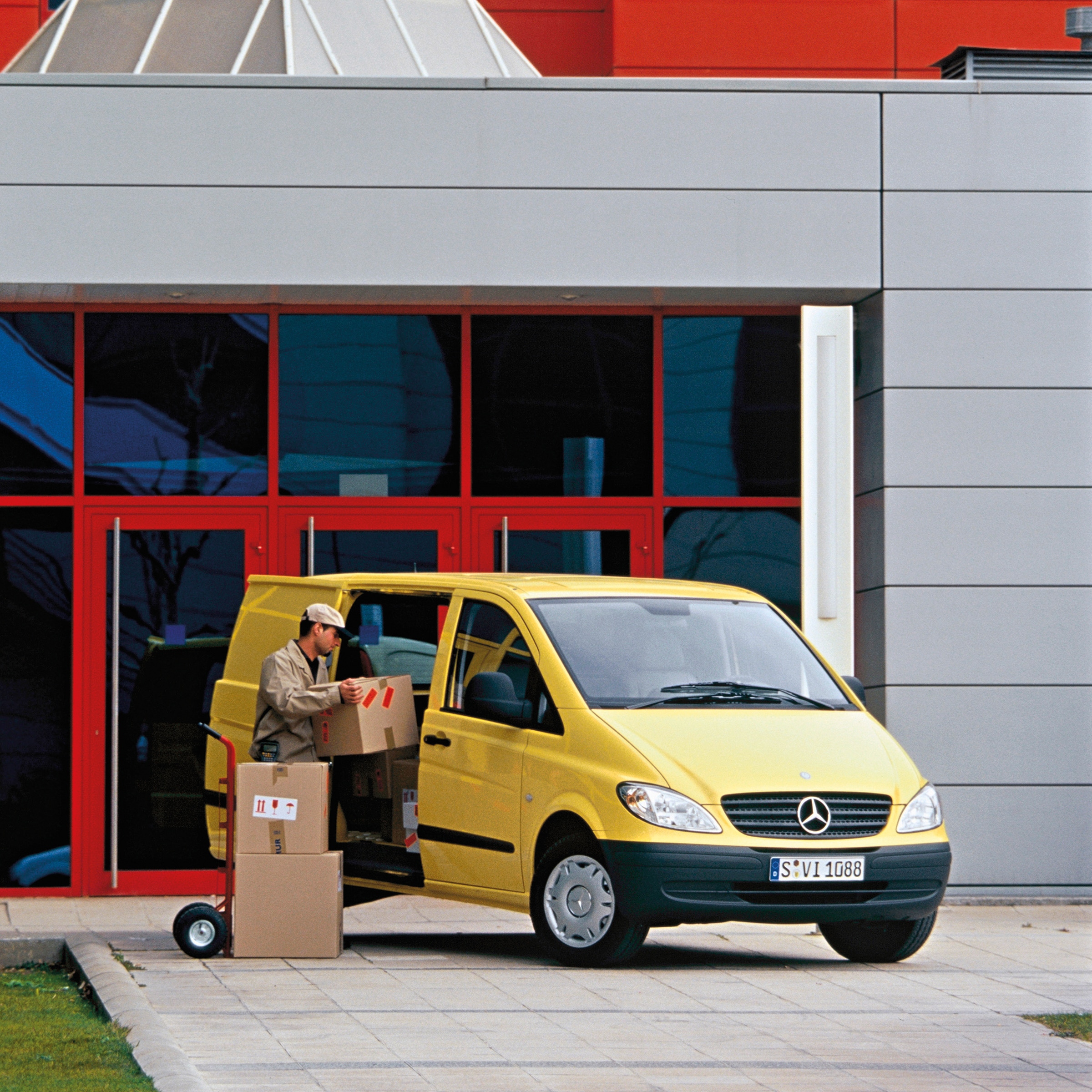 La segunda generación de la Mercedes-Benz Vito | Historia de los vehículos industriales | Mercedes-Benz Un Mercedes-Benz Vito amarillo aparcado frente a un edificio, con un trabajador descargando paquetes